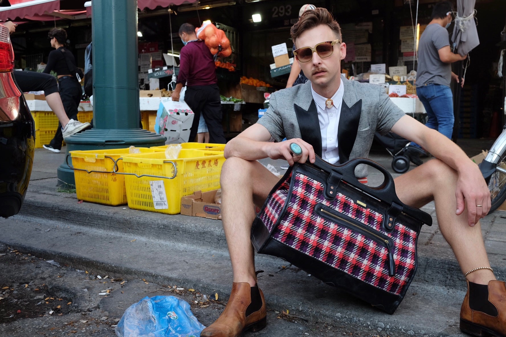 Young man sits on dirty street and shows off plaid duffle bag. 