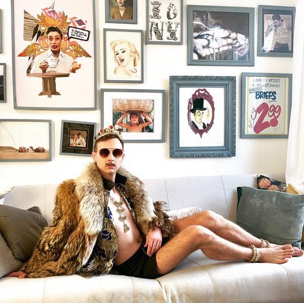 Man in crown and fur coat lounged on the couch with gallery wall behind him 